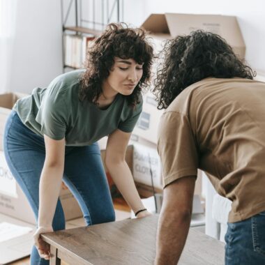 Couple lifting a wooden table while moving into a new home illuminated by daylight.